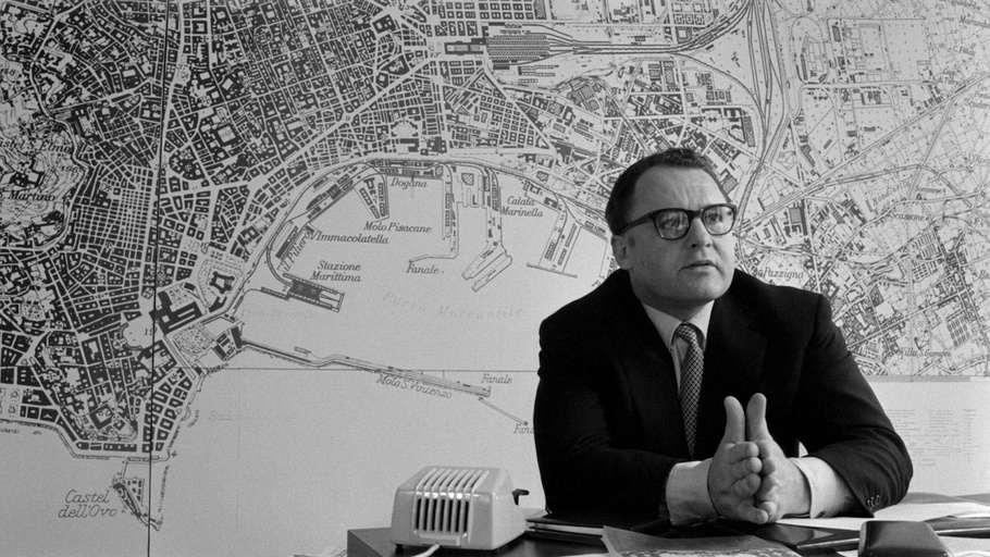 L'attore Rod Steiger in un fotogramma del film: è seduto e si sfrega le mani. Dietro di lui campeggia una cartina della città