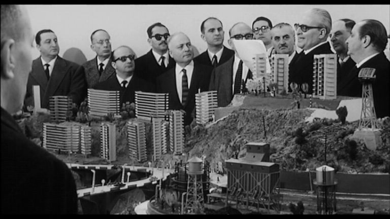 Una scena del film: consiglieri comunali e costruttori, tutti uomini, sorridono. Davanti a loro il modellino degli ecomostri, i palazzi che intendono costruire in città